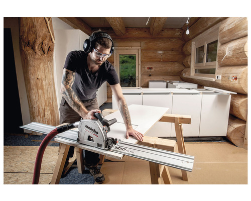 Een man zaagt een werkstuk met een invalcirkelzaag en geleiderail in een blokhut. Hij draagt een veiligheidsbril en gehoorbescherming.