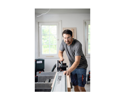 Een man zaagt een houten plank met een handcirkelzaag op een geleiderail, terwijl hij een veiligheidsbril draagt.