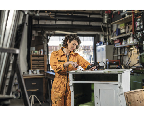 Vrouw verwijdert verf met een heteluchtpistool van een houten kast in een werkplaats.