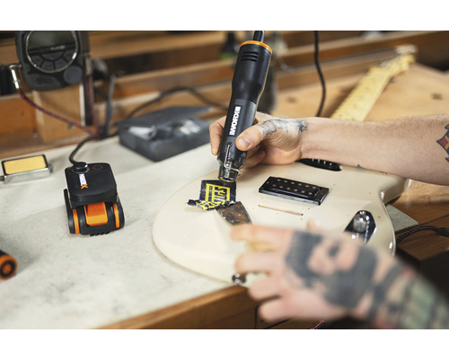 Een man verwijdert met een heteluchtpistool een sticker van een gitaar. Er staat een precisieverhittingsgereedschap op een werkbank.