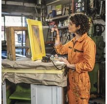 Vrouw spuit verf met een verfspuitpistool op een houten frame in een werkplaats.