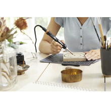 Vrouw werkt met een pyrografiebrander op een stuk hout op een bureau.