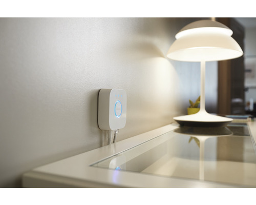 Philips Hue Bridge voor het bedienen van slimme verlichting in huis, aan de muur gemonteerd, naast een tafellamp.