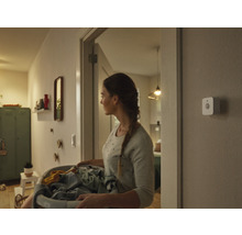 Een vrouw draagt een wasmand door een gang, aan de muur is een smart home bewegingsmelder gemonteerd.