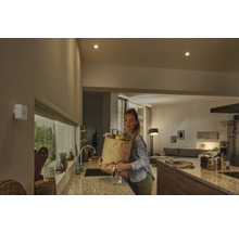 Witte bewegingsmelder aan een muur in een moderne keuken met een vrouw die een boodschappentas draagt.