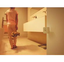 Een witte vierkante bewegingsmelder aan de muur in een zwak verlichte badkamer met een persoon die een teddybeer vasthoudt.