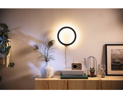 Wandlamp met cirkelvormig ontwerp boven een dressoir versierd met een vaas met pauwenveren, een radio, een cactus en een zandloper.