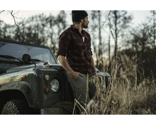 Een man in een geruit hemd leunt tegen een terreinwagen in een landelijke omgeving.
