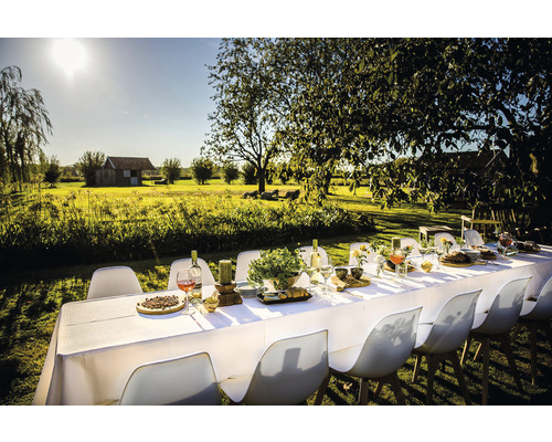 Gedekte lange tafel met stoelen in een tuin, omgeven door bomen en een grasveld.