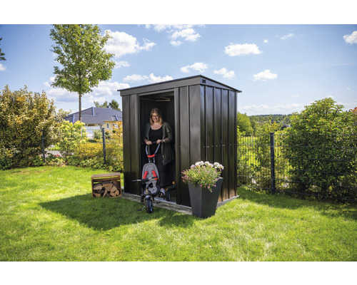 Tuinscene met metalen tuinhuis, vrouw met driewieler, bloembak en stapel hout