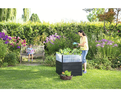 Tuin met verhoogde tuinbak, zittend kind op een bank en een vrouw die in de verhoogde tuinbak werkt