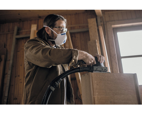 Een vakman schuurt een houten onderdeel met een schuurmachine in een werkplaats, beschermd door een ademhalingsmasker en oogbescherming.