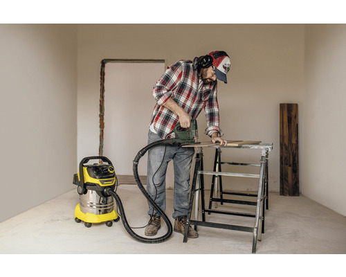 Ambachtsman werkt met een decoupeerzaag aan een werkstuk, een stofzuiger staat klaar om het stof op te zuigen.