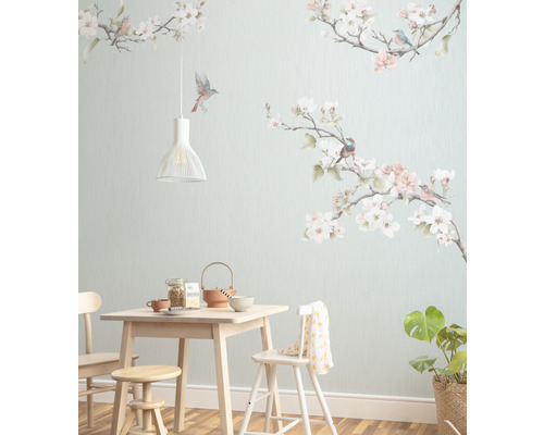 Eetkamer met lichte houten tafel, stoelen, kruk, servies, kamerplant en een muurbehang met vogel- en bloemmotieven.