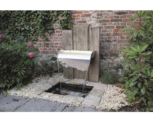 Tuinscène met een roestvrijstalen waterval tegen een bakstenen muur en houten elementen, omgeven door planten en grind