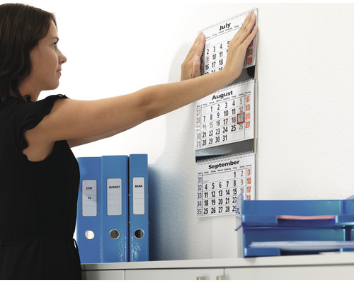 Vrouw bevestigt een kalender aan de muur in het kantoor naast mappen en opbergbakken.