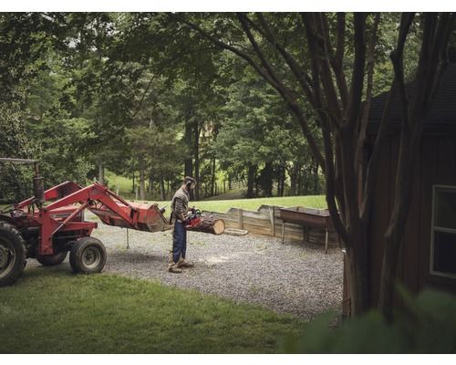 Man zaagt hout met een kettingzaag naast een tractor op een erf.