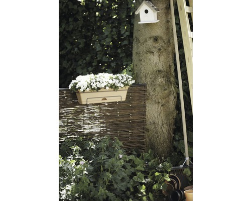 Tuinscene met bloembak, gevlochten schutting, boomstam, vogelhuis en tuingereedschap.