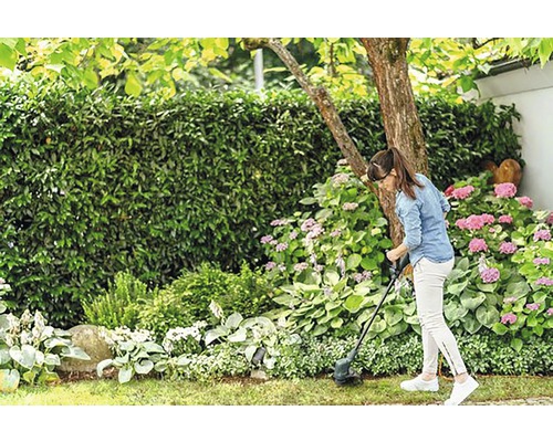Vrouw maait gras met een grastrimmer in de tuin met struiken en bloemen