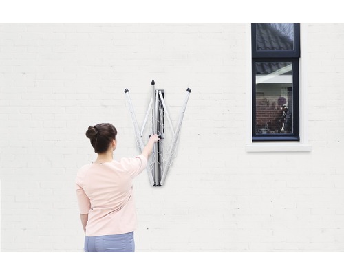 Vrouw bedient een ingeklapt, aan de muur gemonteerd droogrek aan een witte muur naast een raam.