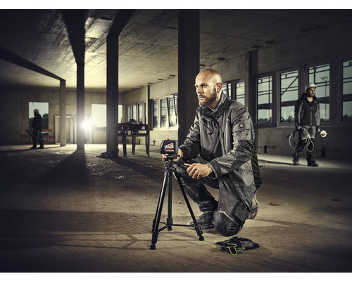 Een arbeider zit gehurkt in een casco en gebruikt een waterpasinstrument op een statief.