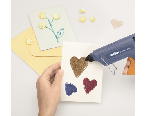 Hand die met een lijmpistool hartjes op een kaart lijmt. Knutselspullen liggen ernaast.