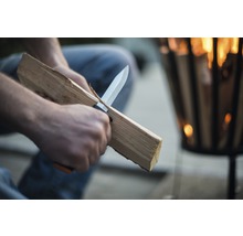 Een persoon snijdt hout met een mes voor een vuurschaal.