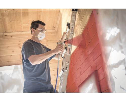 Man met veiligheidsmasker en veiligheidsbril gebruikt een verfspuit op een ladder om een houten muur te schilderen.