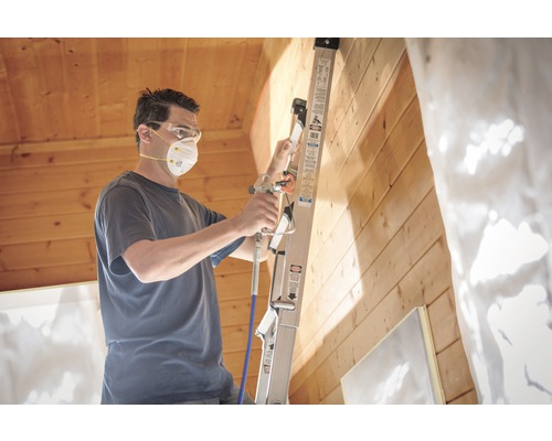 Man met beschermend masker en veiligheidsbril spuitverft een houten muur met een verfspuit vanaf een ladder.