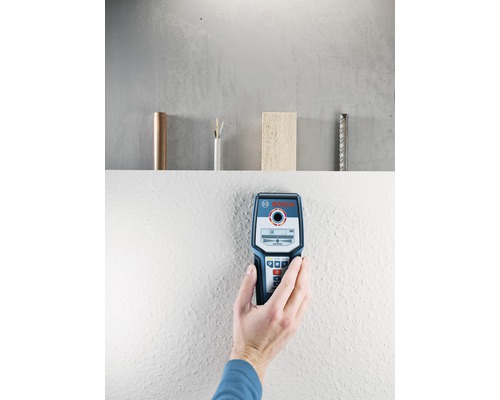 Een hand houdt een Bosch detector tegen een muur. Op een plank op de achtergrond staan een koperen leiding, een elektriciteitskabel, een stuk hout en een wapening.