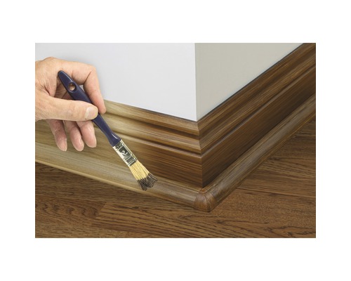 Een persoon verft een bruine plint met een kwast op een muur.