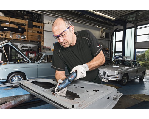 Man werkt met een multifunctioneel gereedschap aan een autodeur in een werkplaats met oldtimers.