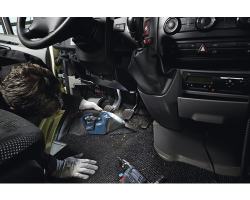 Ambachtsman reinigt voertuiginterieur met snoerloze handstofzuiger. Accuboorschroevendraaier ligt op het tapijt.