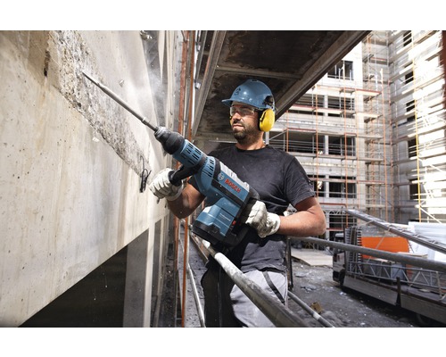 Een bouwvakker met veiligheidshelm en gehoorbescherming werkt met een sloophamer aan een betonnen muur. Bosch Logo.