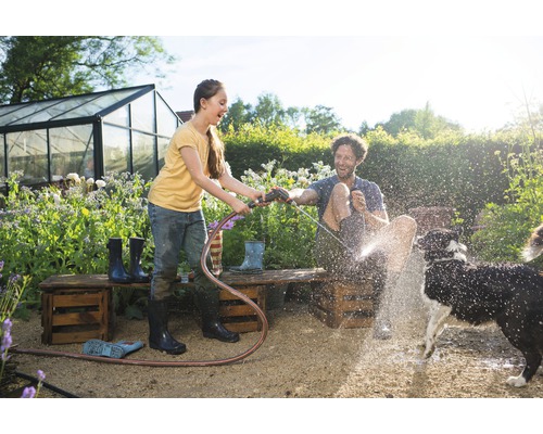 Een jonge vrouw houdt een tuinslang vast en spuit een man en een hond nat in de tuin. Op de achtergrond staat een kas.