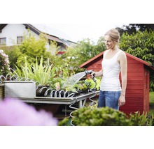 Vrouw besproeit planten met een tuinslang op een plantentafel voor een tuinhuis.