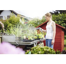 Vrouw bewatert planten met een tuinspuit op een tuintafel voor een rood tuinhuis.