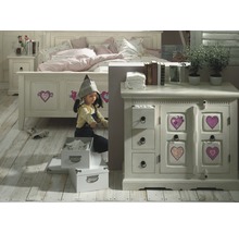 Een meisje speelt in een kinderkamer met bed, nachtkastje en commode in landhuisstijl.