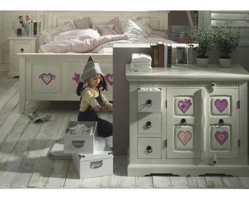 Een meisje speelt in een kinderkamer met bed, nachtkastje en commode in landhuisstijl.