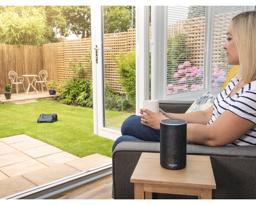 Vrouw met kopje en Amazon Echo naast robotmaaier in de tuin