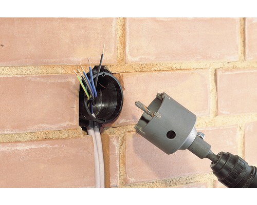 Boormachine met gatenzaag naast een wandcontactdoos in een bakstenen muur