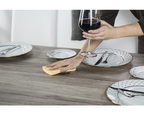 Scene met tafel, couverts en een persoon die een vlek met een spons schoonmaakt terwijl die een wijnglas vasthoudt.