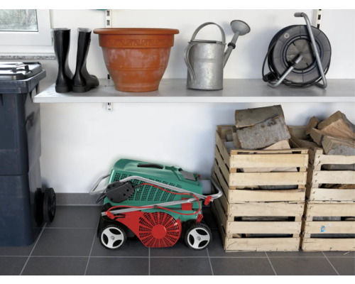 Foto van een garage met planken waarop tuingereedschap zoals rubberlaarzen, een gieter, een bloempot, een grasmaaier en houten kisten zijn opgeslagen.