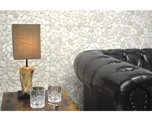 Scene met een tafellamp op een houten tafel, twee glazen, een flesopener en een leren bank voor een muur met kiezelstenen.