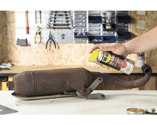 Een man spuit WD-40 roestverwijderaar op een roestige uitlaatpijp in een werkplaats.