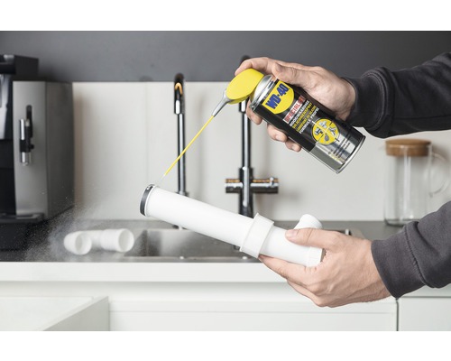 Iemand spuit siliconenspray op een plastic buis onder een wastafel