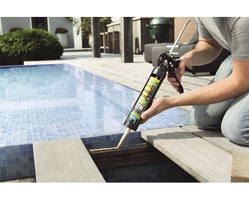 Iemand gebruikt een kitpistool met montagelijm op een houten terras bij een zwembad.