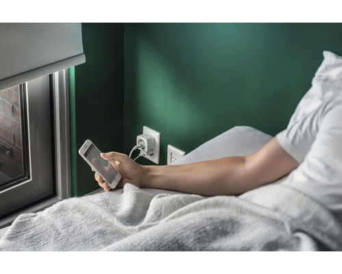 Persoon ligt in bed en houdt een smartphone vast, terwijl een stekkerdoos voor het opladen in de muur zit