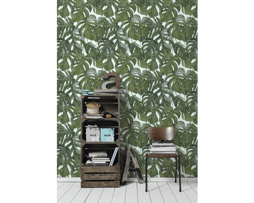 Woonkamer met Monstera-bladbehang, houten krattenkast en stoel met stapel boeken