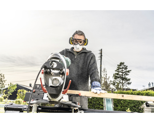 Man met gehoorbescherming, veiligheidsbril en stofmasker werkt met een verstekzaag aan een houten plank